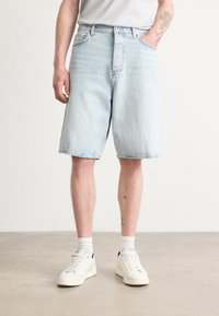Shorts en denim bleu clair avec une coupe ample, dotés de poches avant, d'une fermeture à bouton et d'une finition délavée. Associés à des baskets blanches et des chaussettes.
