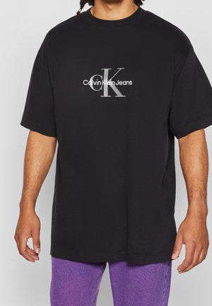 Man draagt een zwart Calvin Klein Jeans T-shirt en paarse broek, staand met armen ontspannen langs het lichaam.