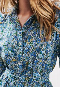 Robe à motif floral présentant un mélange de fleurs bleues, jaunes et vertes sur un fond blanc. Devant à boutons avec une taille ceinturée. Matériau en coton.