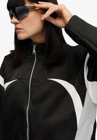 Giacca nera con cerniera e motivi bianchi curvi e colletto alto. Occhiali da sole neri con un accento giallo sull'asta. Tessuto dalla trama liscia.