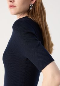 Mørkeblå ribbet strikket topp med korte ermer, med en tettsittende design og subtil tekstur. Modellen har på seg et par sølvfargede hoopøreringer.