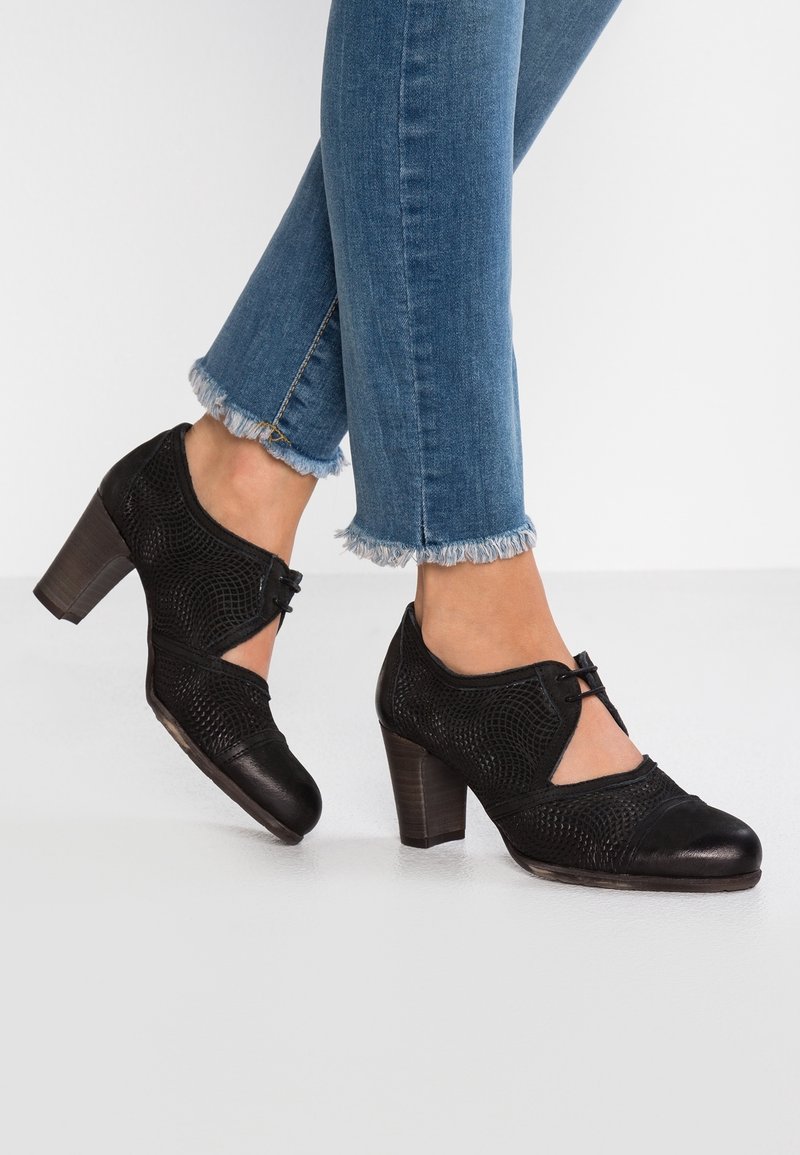 Femme portant des chaussures à talons hauts en cuir noir texturé avec des découpes et des détails en dentelle, associées à un jean bleu à ourlet effiloché.