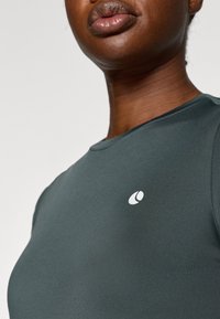 Tamsiai žalia sportinė palaidinė su apvaliu kaklu, pagaminta iš lygaus audinio, su mažai balta logo ant krūtinės.