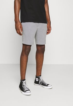 Mężczyzna ubrany w czarno-białe szorty w kratkę, czarną koszulkę i czarne trampki za kostkę z białymi sznurowadłami, na jednolitym tle.