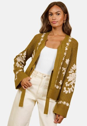 Woman wearing a mustard cardigan with white floral embroidery, a white top, and high-waisted cream pants, posing with one hand on her waist.