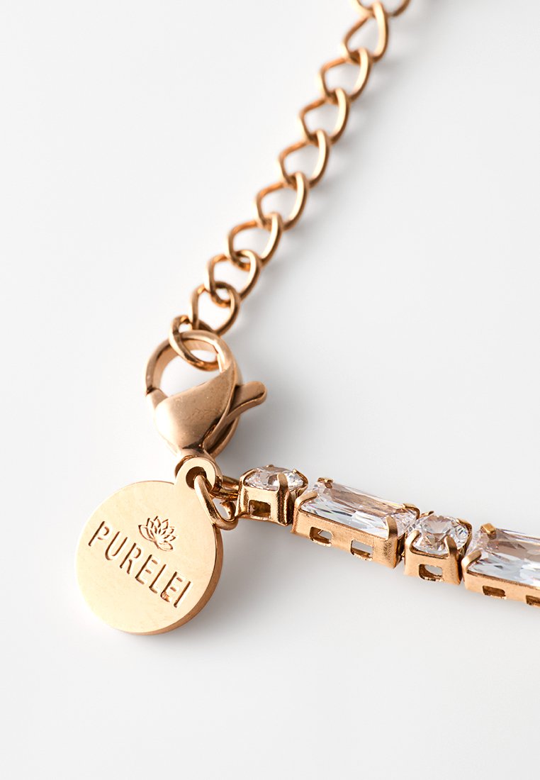 Bracciale in oro rosa con maglie a catena, pietre trasparenti rettangolari e un ciondolo rotondo inciso con "PURELEI." Dettaglio della chiusura visibile.