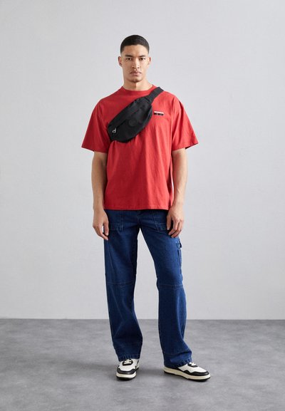 Rød bomulds t-shirt, sort crossbody taske, blå cargo bukser, sort og hvide sneakers. Simpelt design, afslappet pasform og minimal detailering.