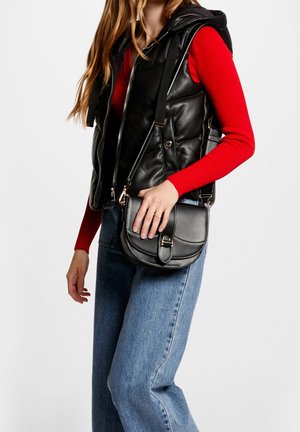 Personne portant un haut rouge à manches longues, une veste matelassée noire, un jean bleu, et tenant un sac en cuir noir porté en bandoulière avec fermeture à boucle.