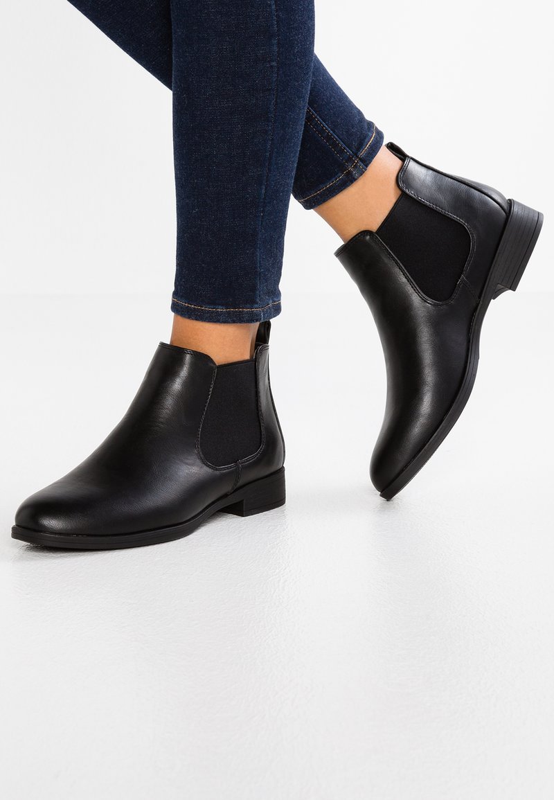 Bottines Chelsea en cuir noir avec des panneaux latéraux en élastique, un petit talon carré et une texture lisse, associées avec un jean noir.