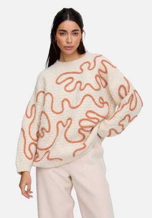 Woman wearing a cream sweater with textured orange squiggle patterns, paired with light beige pants, posing with one hand on her hip.