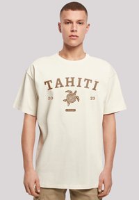 Cream-colored oversized t-shirt featuring a brown graphic print with "TAHITI" and a turtle design. Short sleeves and crew neckline.