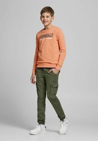 Felpa arancione con testo, pantaloni cargo oliva con tasca laterale, scarpe da ginnastica bianche. Texture morbide, vestibilità casual e colori coordinati.