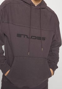 Tmavě fialová mikina s kapucí a přední kapsou, obsahuje vyvýšené černé logo značky, texturovanou tkaninu a zlaté akcenty na šňůrkách.