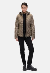 Veste matelassée kaki avec une capuche, fermeture éclair à l'avant et deux poches latérales. Associée à un pantalon noir et des bottes, présentée sur un fond neutre.