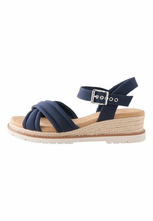 Tmavomodrá klinová sandála s prekríženými látkovými remienkami, prackou na členku, béžovou pletenou medzipodrážkou a bielou gumovou vonkajšou podrážkou.