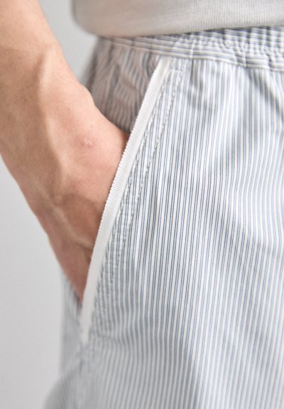 Tissu rayé bleu clair et blanc avec une texture lisse ; une main glissée dans une poche latérale. Couture visible et détail de taille.