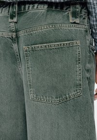 Olivengroene spijkerbroek met een achterzak, voorzien van zichtbare stiksels en een getextureerd denim oppervlak. Elastische tailleband met riemlussen.