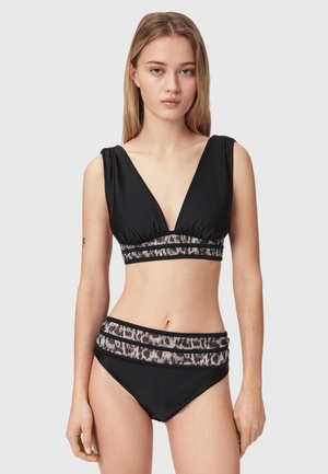 Ung kvinde står iført sort bikini med dyb V-udskæring og dyreprint netbånd øverst og nederst mod en ensfarvet baggrund.