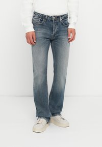 Hombre vestido con jeans rectos desgastados de color azul, suéter de punto blanco y zapatillas blancas, de pie contra un fondo blanco liso.