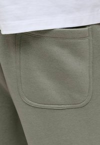 Oliivinvihreät collegehousut, joissa on sileä pinta, suorakaiteen muotoinen tasku ja näkyvät ompelusaumat, yhdistetty valkoiseen yläosaan.