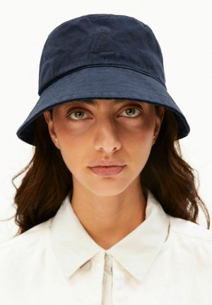 Frau mit hellbraunen Augen und langem dunklem Haar trägt einen dunkelblauen Bucket Hat und ein weißes Hemd mit Kragen, blickt mit neutralem Gesichtsausdruck nach vorne.