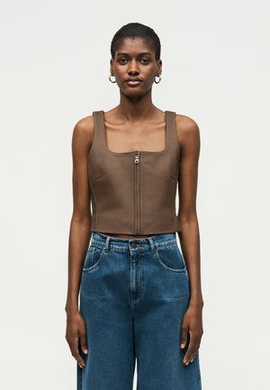 Kobieta o krótkich włosach, nosząca złote kolczyki koła, brązowy, bezrękawnik na zamek oraz niebieskie dżinsy z wysokim stanem, stojąca na jednolitym tle.