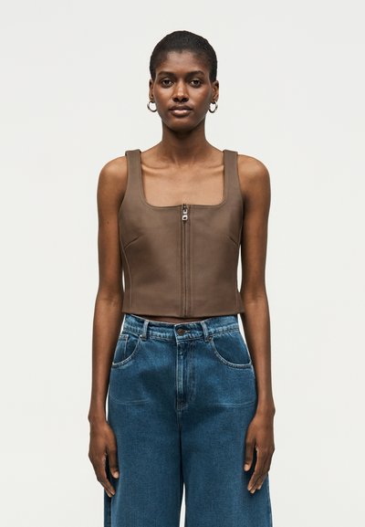 Femme aux cheveux courts portant des boucles d'oreilles créoles dorées, un top sans manches marron zippé et un jean bleu taille haute, debout devant un fond uni.