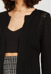 Czarny dziergany kardigan z otwartym przodem, teksturowanym wzorem i prążkowanymi krawędziami, noszony na czarnej bluzce, zestawiony z wzorzystą spódnicą.