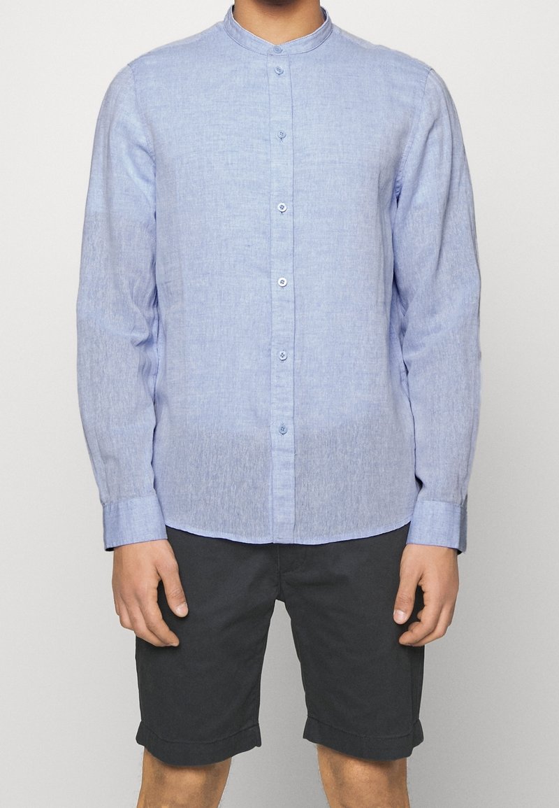 Homme portant une chemise bleu clair à manches longues col mandarin et un short noir retroussé, debout devant un fond uni.