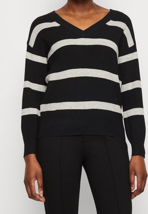 Osoba ubrana w czarny sweter z dekoltem w serek z szerokimi, poziomymi jasnoszarymi paskami oraz czarne spodnie na jednolitym jasnym tle.