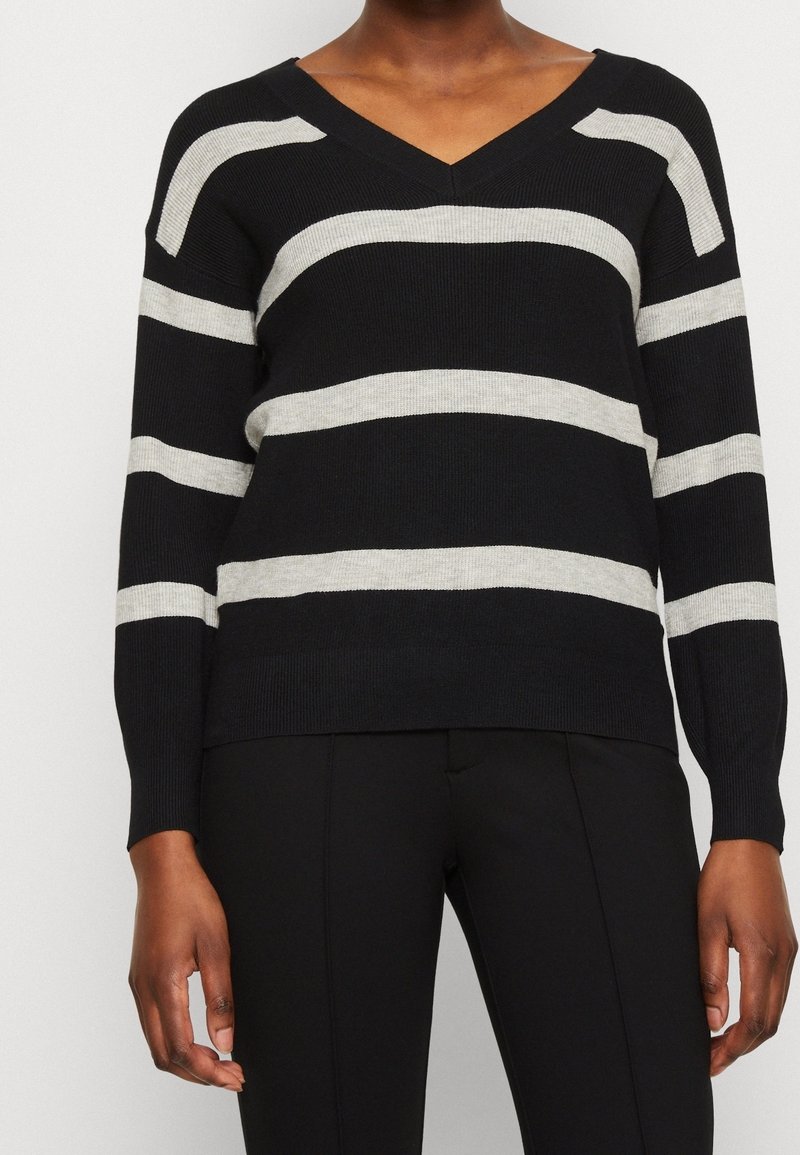 Personne portant un pull noir à col en V avec de larges rayures horizontales gris clair et un pantalon noir sur un fond uni clair.