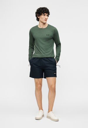 Mees lokkis juustega, kes kannab rohelist pika varrukaga särki, musti lühikesi pükse logoga ning valgeid tossusid, seisab käed taskutes.