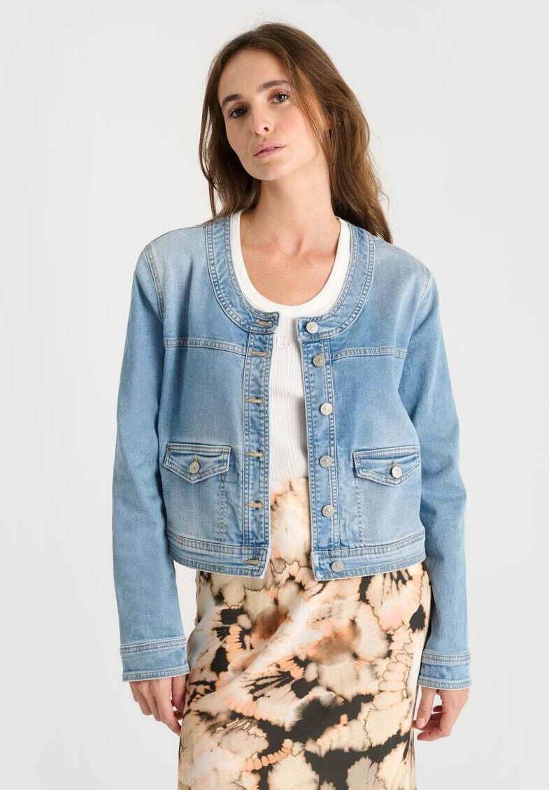 Lichtblauwe denimjas met een ronde halslijn, een knoopsluiting aan de voorkant, twee zakken onderaan en lange mouwen. Draagt over een gestreepte rok.