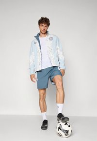 Helehall sinine jakk lillekujundusega, valge t-särk, sinised lühikesed püksid ja mustad jalanõud. Jalg palliga jalgpalliplatsil.