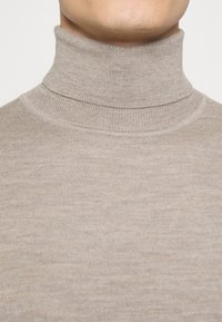 Gros plan sur une personne portant un pull beige à col roulé côtelé, montrant le cou et la partie supérieure de la poitrine.