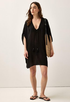 Kvinde iført løs sort kjole, bærende en flettet skuldertaske, stående barfodet i sorte sandaler mod en ensfarvet baggrund.