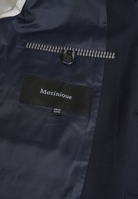 Innenansicht eines marineblauen Blazers mit einer gestreiften Tasche, ausgestattet mit einem schwarzen Etikett, auf dem "Matinique" steht, sowie Pflegeetiketten in Weiß. Glattes, elegantes Material.