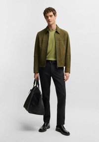 Green suede jacket with a zip, layered over a green top. Black trousers and polished black shoes. Holding a large black bag.