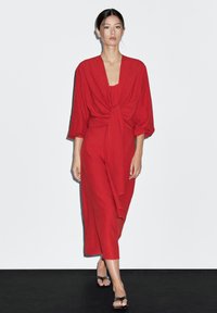 Červené midi šaty s krátk�ými širokými rukávy a uzlovým detailem na přední části. Hladká tkanina a volný střih, sahající těsně pod kolena.