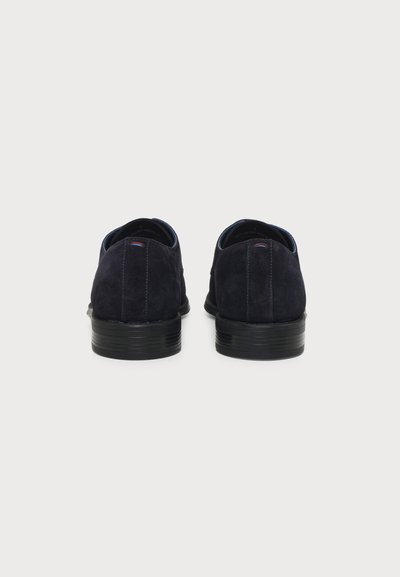 Paire de chaussures habillées en daim noir vues de dos avec des talons noirs empilés et une couture rouge et blanche subtile près du dessus.