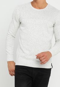 Maglione grigio chiaro lavorato a maglia con texture a costine sulle spalle, collo arrotondato e polsini aderenti, abbinato a jeans scuri.