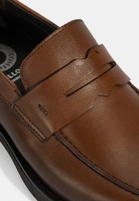 Bruine leren instapper met een ronde neus, decoratieve uitsparingen en contrasterende stiksels. Beschikt over een gestructureerd oppervlak en een zwarte rubberen zool.