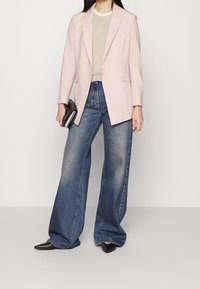 Blazer rose clair aux épaules structurées, associé à un haut beige et un jean bleu délavé à jambes larges. Chaussures noires à bout pointu et pochette.