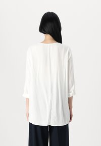 Donna con i capelli lunghi neri che indossa una blusa bianca a maniche lunghe ampia e pantaloni neri a gamba larga, vista di spalle su uno sfondo semplice.