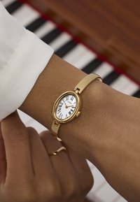 Gouden ovale polshorloge met Romeinse cijfers op een witte wijzerplaat, verbonden met een gladde gouden band. Een hand rust op een pianotoetsenbord.