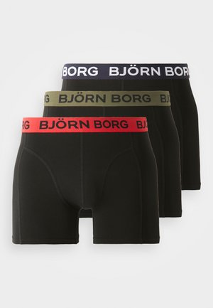 Trīs melnu bokseru bikses pāri ar kontrastējošām jostām: sarkana, olīvu zaļa un jūras zila, ar logotipa tekstu un pieguļošu dizainu.