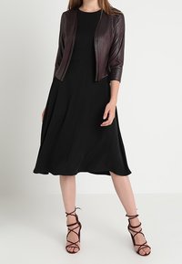 Robe noire avec une jupe fluide, associée à une veste en cuir bordeaux foncé ajustée. Des sandales à talons hauts marron à brides complètent le look.