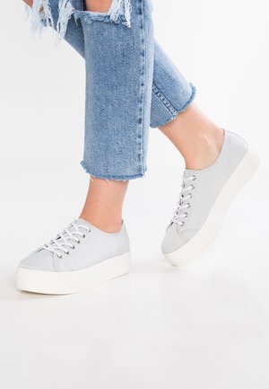 Nogi w krótkich, postrzępionych, jasnoniebieskich dżinsach i jasnoszarych sneakersach na platformie z białymi sznurowadłami na białym tle.