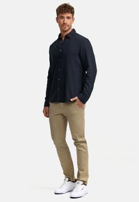 Camisa de botones azul marino, con un corte holgado y mangas largas, combinada con pantalones beige y zapatillas blancas, de pie frente a un fondo liso.