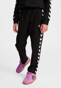 Zwarte sportbroek met witte zijkanten in chevronpatroon, gecombineerd met paarse sneakers met donkere accenten. Soepele stof en een ontspannen pasvorm.